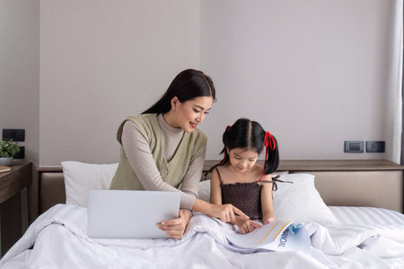 A young woman is working from home with her adorable daughter, watching work together and happily teasing each other in the bedroom.の写真素材