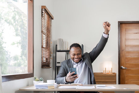 Young successful African American businessman uses laptop to work on white wooden table, expressing excitement in office.の写真素材