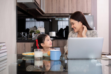 a single mother, sits at home working on a laptop with her daughter beside her watching and encouraging her.の写真素材