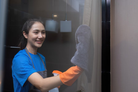 A beautiful housekeeper wears orange gloves and uses a cloth to clean the glass of the room.の写真素材