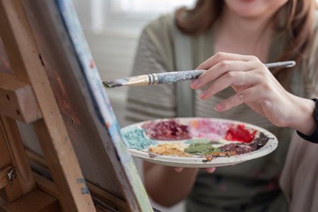 Young Asian artist woman holding a palette of colorful paints is intently painting acrylic paints on canvas in a painting studio.の写真素材