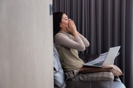 A middle-aged woman is tired while working from home in her bedroom with her laptop and pile of...