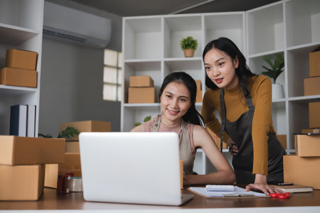 Two women working on laptop and packing boxes in warehouseの写真素材