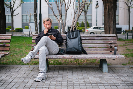 Man sitting on a park bench using smartphone with backpack and camera. Concept of urban relaxation...