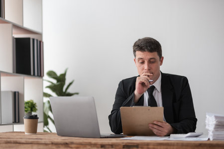 Young Businessman Disappointed at Work, Reflecting on Challenges, Sitting at Desk with Laptop and Clipboard in Modern Office Settingの写真素材