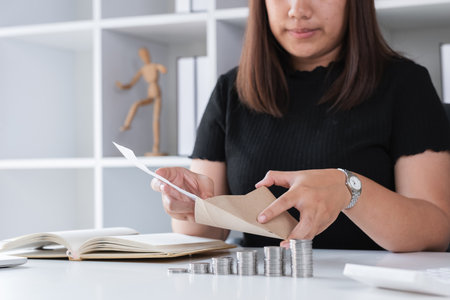 Young Woman Planning to Save Money by Counting Coins and Organizing Finances at Home Office Desk with Notebook and Envelopes, Concept of Financial Planning and Budgeting for Future Goalsの写真素材