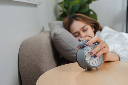 Young Woman Turning Off Alarm Clock in the Morning, Starting Her Day with a Relaxed Wake-Up Routine, Cozy Home Environmentの写真素材