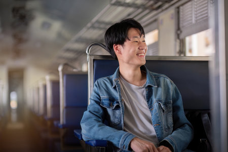 Young Man Sitting on Train, Smiling and Looking Out the Window, Casual Denim Jacket, Relaxed Travel Vibeの写真素材
