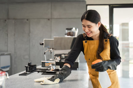 Female Employee Cleaning Coffee Shop Counter with Spray Bottle and Cloth in Modern Cafe Settingの写真素材