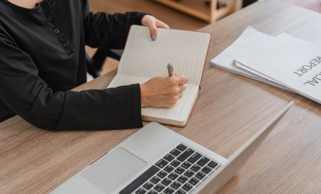 Woman Working from Home Writing in Notebook with Laptop on Wooden Desk in Modern Home Office Settingの写真素材