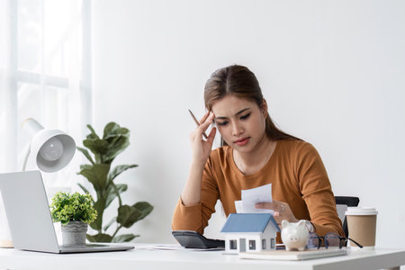 Young Woman Stressed While Selling a House, Working at Desk with Laptop, Documents, and Model Houseの写真素材