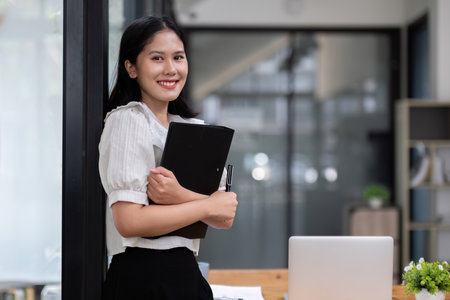 Confident Business Woman Holding Documents in Modern Office Setting, Smiling and Ready for Successの写真素材