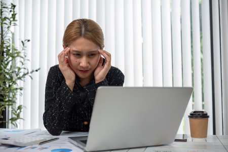 Stressed Business Woman Working on Laptop in Office with Coffee and Documents, Feeling Overwhelmed and Anxiousの写真素材