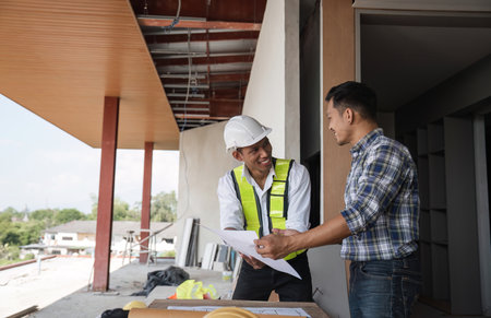 Engineer Discussing Construction Plans with Colleague on Site, Holding Blueprint, Planning Work, Wearing Safety Gearの写真素材