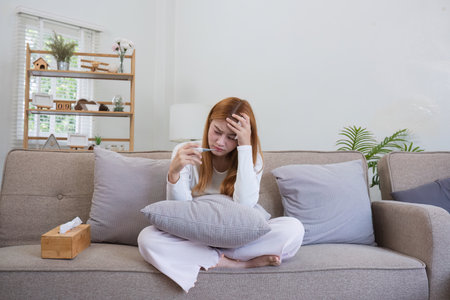 Young Woman Checking Her Temperature with a Thermometer While Sitting on a Sofa at Home