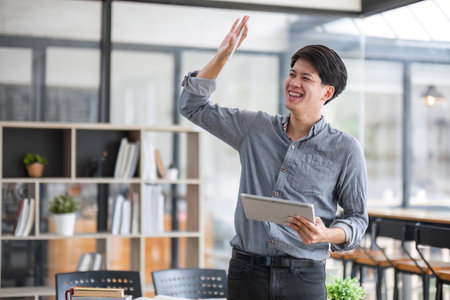 Young Office Worker Smiling and Waving While Holding a Tablet in a Modern Office Environmentの写真素材