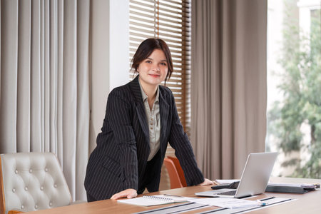 Confident Businesswoman in Modern Office Setting with Laptop and Documents on Desk, Professional...
