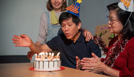 A man is blowing out candles on a birthday cake while two women watch