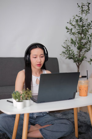 Young Woman Studying Online from Home with Laptop and Headphones, Sitting on Floor in Modern Living...