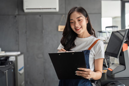 Young Barista Managing a Modern Coffee Shop with a Smile, Holding a Clipboard and Wearing an Apron,...
