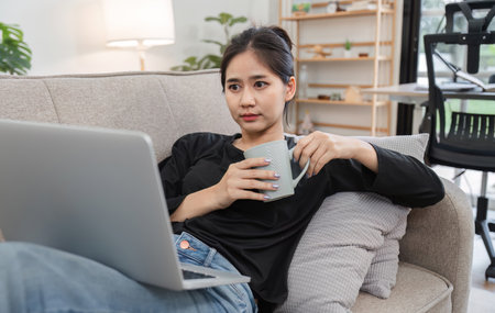 Young Accountant Working on Financial Documents at Home, Analyzing Data on Laptop While Relaxing on Couch with Coffeeの写真素材