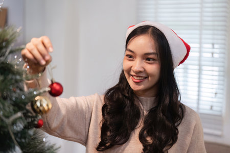 Young Woman Joyfully Decorating a Christmas Tree with Ornaments in a Cozy Home Setting During the Festive Holiday Seasonの写真素材