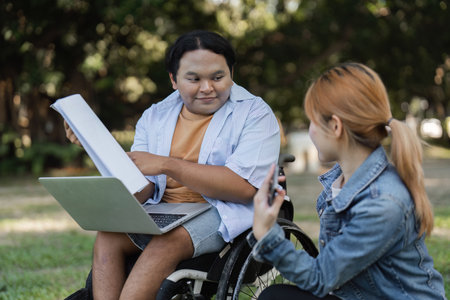Young Man in Wheelchair Collaborates with Friend on Paperwork Outdoors in a Park Settingの写真素材