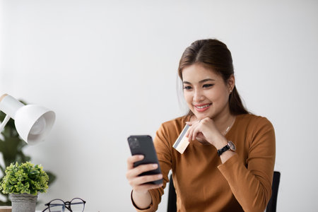 Woman Shopping Online Holding a Credit Card and Smartphone in a Modern Home Office Settingの写真素材