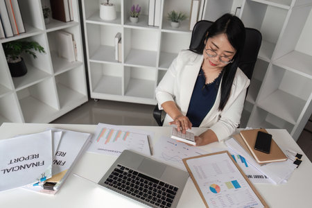 Middle-aged Female Accountant Working on Financial Documents in Modern Office Setting with Laptop...