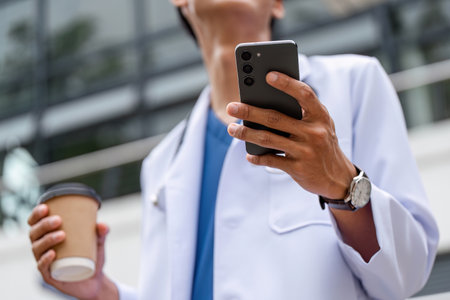 Male Doctor in Professional Uniform Holding Coffee and Smartphone Outdoors in Urban Settingの写真素材