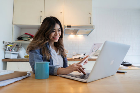 Young Freelance Woman Working from Home in the Kitchen with Laptop and Coffee, Smiling and Reviewing Documentsの写真素材