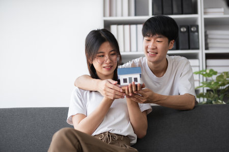 Young Couple Exploring Real Estate Opportunities, Holding a Model House, and Discussing Future Home Purchase Plans in a Modern Living Room Settingの写真素材