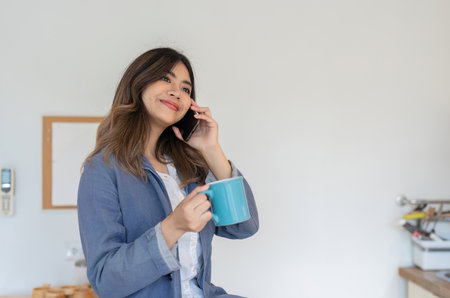 Young Freelance Woman Working from Home, Talking on Phone in Dining Room, Holding Coffee Mug, Remote Work Conceptの写真素材