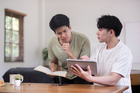 Young Gay Couple Sitting Together at Home, Engaging in a Discussion with a Book and Tabletの写真素材