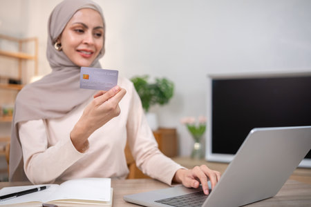 A young Muslim woman wearing a hijab sits contentedly shopping on her laptop, paying through an online banking app and holding a credit card.の写真素材