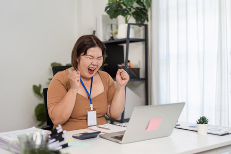 Joyful Female Accountant Celebrates Professional Success in Modern Office Setting with Laptop and Documentsの写真素材