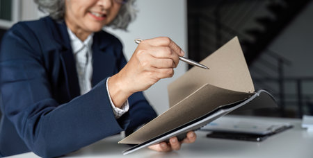 Elderly Businesswoman Engaged in Reading a Book at Her Desk in a Modern Office Settingの写真素材