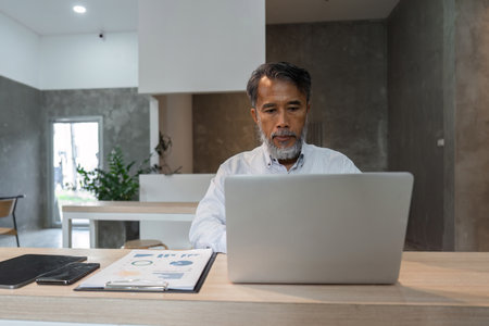 Concentrated male businessman reviewing data on laptop with documents and smartphone in a modern...