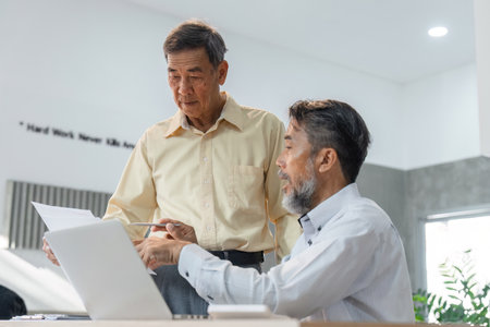 Seasoned businessmen discussing project updates while examining documents in a bright office environmentの写真素材