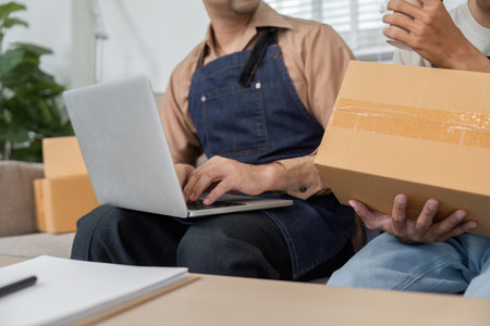 Delivery professional typing on a laptop while another holds a package in a collaborative workspaceの写真素材