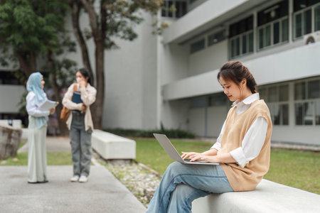 Multicultural woman studying on laptop outdoors while friends converse in university setting.の写真素材