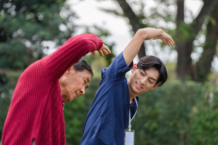 Young caregiver encouraging elderly man to perform arm stretching exercise in gardenの写真素材