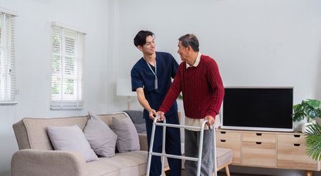 Caring nurse smiling while assisting elderly man with walker in sunny living roomの写真素材