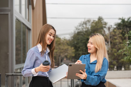 Two diverse women discussing ideas with a tablet and coffee outdoors in a modern environment.の写真素材