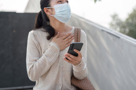 Young woman wearing a mask, holding smartphone, and showing emotion outdoors.の写真素材