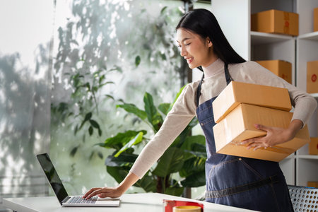 Young woman in apron carrying boxes while using a laptop in a modern workspace.の写真素材