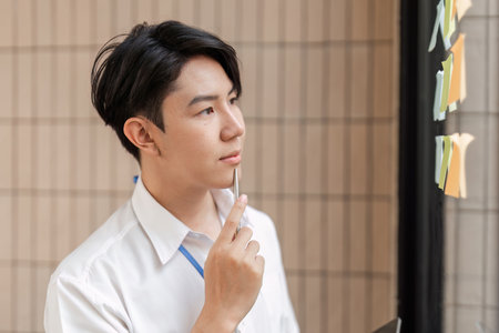 Young professional contemplating sticky notes on glass wall, strategizing tasks and improving workflow in modern office.の写真素材