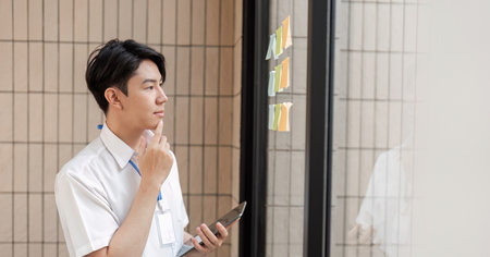 Young Asian male office worker thoughtfully analyzing sticky notes on glass wall in modern workspace, focusing on planning and strategy.の写真素材