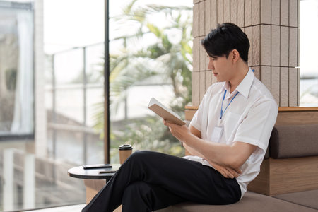 Young Asian male office worker reading book in modern workspace, enjoying leisure time and personal development.の写真素材