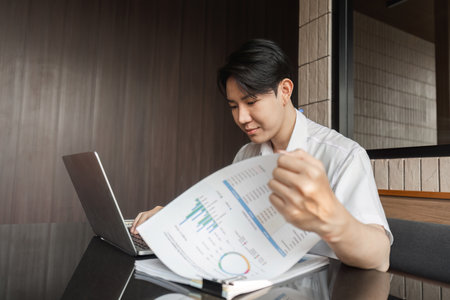 Young professional analyzing financial report while working on laptop in modern office environment.